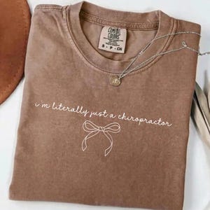 Puede incluir: Una camiseta marrón Comfort Colors con el texto "i'm literally just a chiropractor" y un lazo. Un collar plateado con un colgante está sobre la camiseta. La camiseta está doblada.
