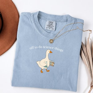 May include: A light blue t-shirt featuring a graphic of a white goose carrying a test tube rack and a flask. The text "off to do science things" is printed above the goose. The shirt has a Comfort Colors label indicating size S and 100% cotton.