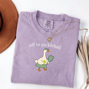 May include: A light purple t-shirt featuring a graphic of a white goose wearing a green visor and skirt, holding a pickleball paddle and ball. The text above the goose reads off to pickleball. A gold chain necklace rests on the shirt collar.