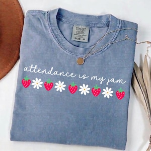 Könnte beinhalten: Blaues Comfort Colors T-Shirt mit dem Schriftzug "attendance is my jam" in weißer Schrift. Das Design umfasst rote Erdbeeren und weiße Gänseblümchen. Eine silberne Halskette mit goldenem Anhänger liegt auf dem Shirt.