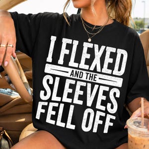 May include: A black short-sleeve t-shirt featuring white distressed-style text that reads I FLEXED AND THE SLEEVES FELL OFF. The shirt has a crew neckline and is worn with a layered gold-tone necklace featuring a small circular pendant.