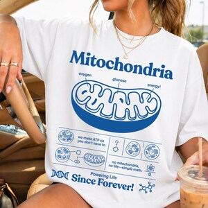 Puede incluir: Camiseta blanca con un gráfico azul de un diagrama de mitocondrias y la palabra "Mitochondria" en azul. La camiseta también tiene el texto "Powering Life Since Forever!"