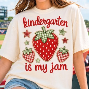 May include: A cream-colored t-shirt featuring a graphic with the red text "kindergarten is my jam" and three strawberries with green and white gingham leaves. The design includes small red and green gingham stars surrounding the strawberries.