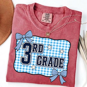 Könnte beinhalten: Ein altrosa T-Shirt mit einem Grafikdruck auf hellblau-weiß kariertem Hintergrund, dunkelblauem Schriftzug 3rd GRADE, zwei hellblauen Schleifen und kleinen weißen Sternen. Das T-Shirt hat einen Rundhalsausschnitt und ein Comfort Colors Etikett.