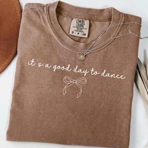 May include: A brown Comfort Colors t-shirt with the phrase "it's a good day to dance" in white script, along with a bow graphic. A silver necklace with a pendant is draped across the shirt.