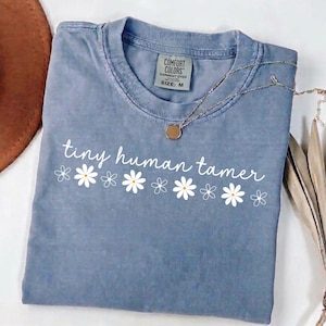 Op de afbeelding: Een blauw Comfort Colors T-shirt met de tekst "tiny human tamer" en madeliefjesafbeeldingen. Het shirt heeft een ronde hals en is maat M. Een zilveren ketting met een ronde hanger ligt over het shirt.