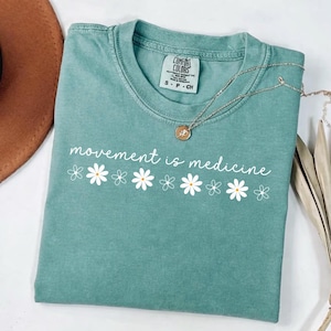 Puede incluir: Una camiseta Comfort Colors de color verde azulado con la frase "movement is medicine" en escritura blanca, acentuada con gr&aacute;ficos de margaritas blancas. Un collar plateado con un colgante descansa sobre la camiseta doblada.