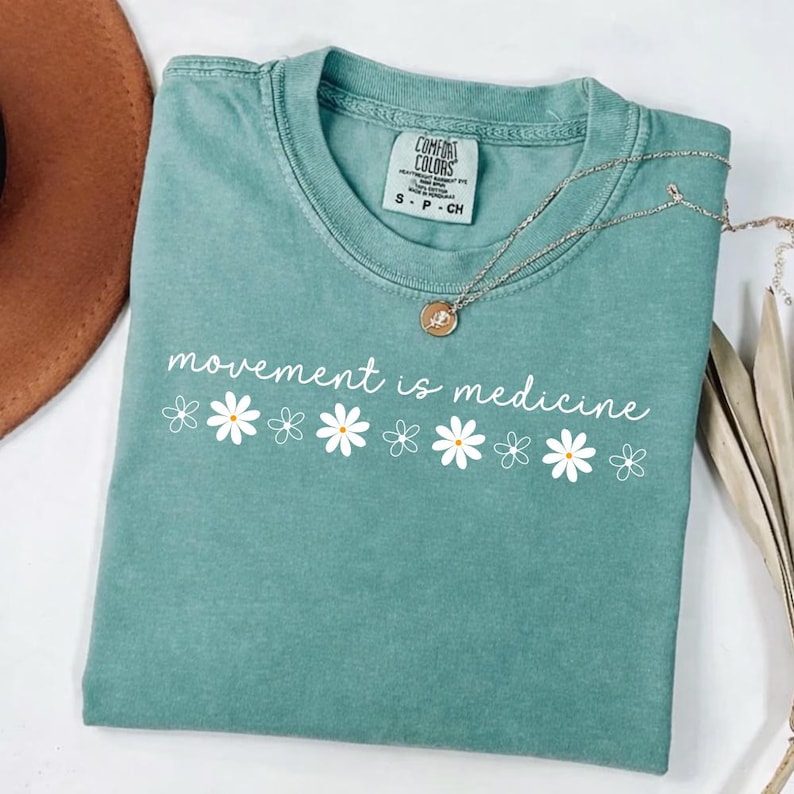 Puede incluir: Una camiseta Comfort Colors de color verde azulado con la frase "movement is medicine" en escritura blanca, acentuada con gr&aacute;ficos de margaritas blancas. Un collar plateado con un colgante descansa sobre la camiseta doblada.