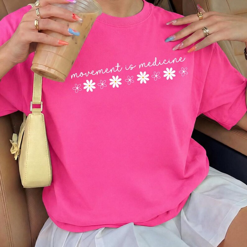 Puede incluir: Una camiseta rosa brillante con el texto blanco "movement is medicine" y gr&aacute;ficos de margaritas blancas. La camiseta de manga corta parece estar hecha de un material suave. Tambi&eacute;n se ve un bolso de mano amarillo claro.