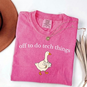 May include: A pink short-sleeved t-shirt with a graphic of a white goose wearing a stethoscope and holding a coffee cup and a clipboard. The text "off to do tech things" is printed in white above the goose. The shirt has a Comfort Colors label.