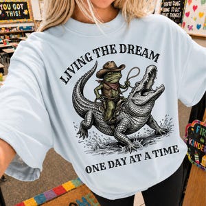 Op de afbeelding: Lichtblauw T-shirt met een afbeelding van een kikker-cowboy die op een alligator rijdt, met de tekst "LIVING THE DREAM ONE DAY AT A TIME". De kikker draagt een cowboyhoed en vest. Het ontwerp is in zwarte inkt.