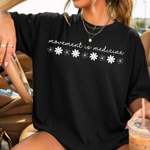 Puede incluir: Camiseta negra con la frase "movement is medicine" en blanco y ilustraciones de margaritas blancas. La camiseta es de estilo casual, adecuada para el uso diario.