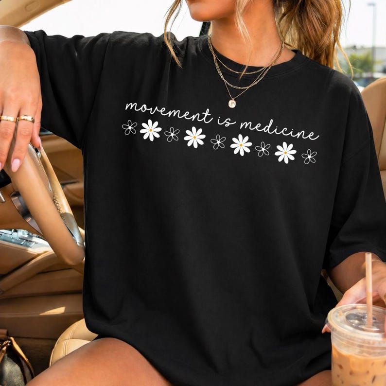 Puede incluir: Camiseta negra con la frase "movement is medicine" en blanco y ilustraciones de margaritas blancas. La camiseta es de estilo casual, adecuada para el uso diario.