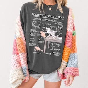 May include: Dark gray long-sleeve shirt with the text "WHAT CATS REALLY THINK" and a diagram of a cat experiment. The experiment includes a cat, a vase, and mathematical formulas. A colorful, striped cardigan is worn over the shirt.