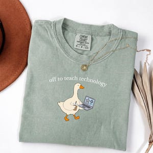 May include: A sage green t-shirt featuring a graphic of a white goose holding a coffee cup and a laptop. The text "off to teach technology" is printed above the goose. The shirt has a crew neckline and a Comfort Colors brand label inside.