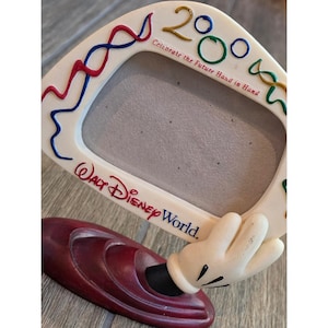 Może przedstawiać: Kremowa ramka na zdjęcia Walt Disney World z napisem 2000 oraz Celebrate the Future Hand in Hand. Ramka jest ozdobiona czerwonymi, niebieskimi i zielonymi falistymi liniami i wsparta na podstawie z białą dłonią w rękawiczce z kreskówki.
