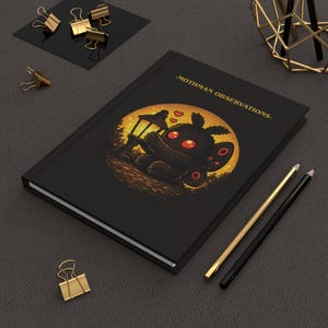 May include: Black journal with the text "-MOTHMAN OBSERVATIONS-" and an illustration of a cartoon moth holding a lamp. The moth has red eyes and is set against a yellow background. Gold and black pencils and gold binder clips are also visible.