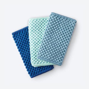 May include: Three rectangular washcloths in shades of blue and green are arranged on a white background. The washcloths have a textured, raised dot pattern. The colors include deep blue, light blue, and a soft green.
