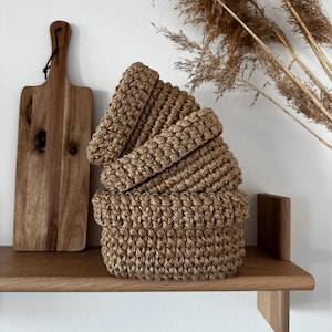 Puede incluir: Tres cestas marrones apiladas y tejidas con un borde superior enrollado, sobre una estantería de madera. Una tabla de cortar de madera cuelga de la pared detrás de las cestas. La hierba ornamental seca añade un elemento natural.