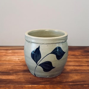 May include: A cylindrical stoneware crock with a light beige, speckled finish and a thick, rounded rim. The exterior features a hand-painted design of a dark blue leafy branch with three distinct leaves extending from a single thin stem.