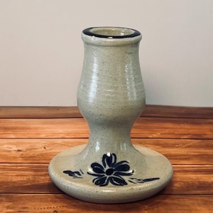 May include: A speckled light gray ceramic candlestick holder with a wide circular base and a flared top rim. The piece features a hand-painted dark blue flower design on the base and a dark blue band around the top opening of the candle holder.