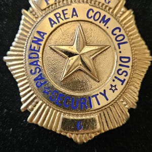 May include: A gold-toned metal badge shaped like a shield with a central star. The text reads PARKING AREA COM. COL. DIST. PASADENA SECURITY with the number 6 at the bottom. The badge features a textured border and blue lettering throughout.