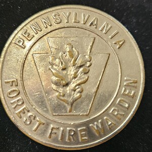 Puede incluir: Una insignia circular de metal dorado con el texto PENNSYLVANIA en la parte superior y FOREST FIRE WARDEN en la inferior. En el centro, una forma de clave de bóveda en relieve contiene un diseño de hoja de roble, todo en acabado metálico.
