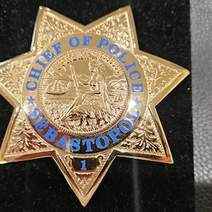 May include: A gold-coloured seven-pointed star badge featuring an embossed central seal with a figure and landscape. The blue text reads CHIEF OF POLICE SEBASTOPOL with the number 1 at the bottom point. The badge has intricate engraved details.