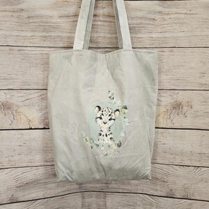 May include: A light gray tote bag featuring a central illustration of a spotted leopard cub inside a soft green oval frame, accented with delicate green eucalyptus leaves surrounding the base of the portrait and two long shoulder straps.
