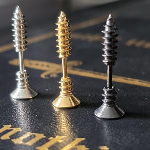 Peut inclure: Trois boucles d'oreilles clous en forme de vis, argentées, dorées et noires. Les boucles d'oreilles ont une tête de vis avec une tige et une base évasée. Les boucles d'oreilles sont présentées sur une surface sombre.