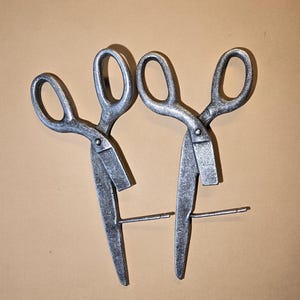 May include: Two vintage metal scissors with oval finger holes, set against a tan backdrop. The scissors have a silver-grey finish and a simple design. The blades are open, and a metal rod connects them.