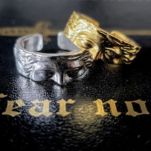 Puede incluir: Dos anillos abiertos, uno en tono plateado y otro en tono dorado, cada uno con un rostro humano esculpido con ojos cerrados y detalles de cabello fluido. Los anillos están colocados sobre una superficie negra con el texto dorado fear not.
