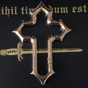 May include: A metallic, rose gold-coloured cross-shaped carabiner with a spring-loaded gate mechanism. The carabiner features a pointed, ornate edge design and is positioned over a black surface with the text "nihil timendum est" and "fear nothing".