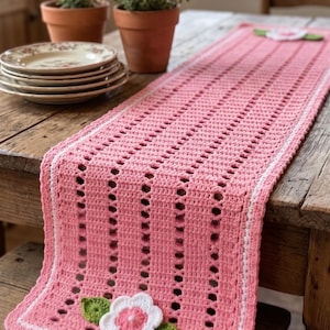 Könnte beinhalten: Ein rosa gehäkelter Tischläufer mit weißem Rand und dekorativen Blumenakzenten. Der Läufer hat ein offenes Design mit vertikalen Lochreihen. Teller und Topfpflanzen befinden sich im Hintergrund.