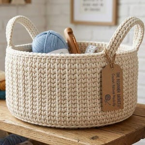 Może przedstawiać: Kremowy, szydełkowy kosz z dwoma uchwytami, wypełniony niebieską przędzą, drewnianym szydełkiem i miarką. Etykieta głosi "The Beginner Basket // Crochet Pattern Mock-Up."