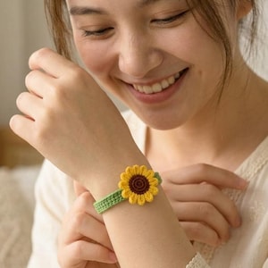 以下が含まれることがあります： 黄色と茶色のヒマワリのデザインが特徴的な、手作りのかぎ針編みブレスレット。 ヒマワリが焦点で、緑色のバンドにセットされています。 ブレスレットは魅力的なアクセサリーです。