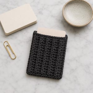 May include: A dark charcoal gray rectangular crochet card holder featuring a vertical ribbed texture. The holder is constructed with a thick yarn and contains a single blank off-white card tucked inside the top opening of the sleeve.