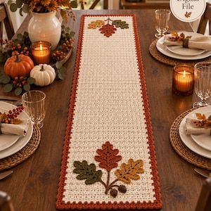 Puede incluir: Un camino de mesa rectangular de ganchillo en color crema con un borde color óxido, con hojas de otoño aplicadas en verde, naranja y amarillo con pequeñas bellotas marrones en los extremos. El texto Digital File es visible arriba a la derecha.