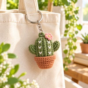 Puede incluir: Un llavero de cactus de ganchillo que presenta un cactus verde con detalles bordados en forma de V blanca, una pequeña flor rosa con centro amarillo y una maceta marrón, todo unido a una anilla y cadena de metal plateado en un bolso de lona.