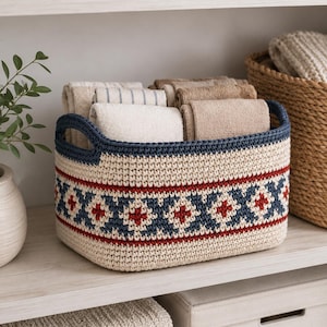 May include: A rectangular crochet basket featuring a cream-colored base with a dark blue rim and handles. The exterior is decorated with a horizontal band of dark blue and red geometric star patterns on a cream background, filled with folded towels.