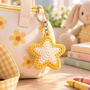 Peut inclure: Un porte-clés étoile en crochet avec une bordure extérieure jaune et un centre blanc, attaché à un anneau en métal argenté. Le porte-clés est suspendu à un sac en tissu crème orné de motifs de fleurs jaunes et d'une sangle à carreaux.