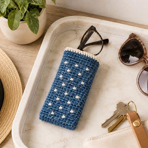 May include: A rectangular crochet glasses case in deep blue with a repeating pattern of small white star-like stitches and a cream-colored top border. The case holds a pair of dark-rimmed eyeglasses, with a pair of sunglasses and keys nearby.