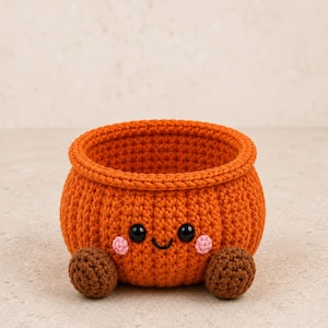 Puede incluir: Una cesta redonda de ganchillo naranja con una cara sonriente, ojos de plástico negro, una boca bordada y pequeñas mejillas rosas. La cesta tiene dos pies esféricos y texturizados de color marrón en la base frontal, formando un cuenco.