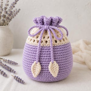 May include: A round, crocheted drawstring pouch featuring a lavender-colored base and a cream-colored upper section with an open-work pattern. The pouch is cinched with a matching lavender cord that ends in two small, cream-colored leaf-shaped accents.