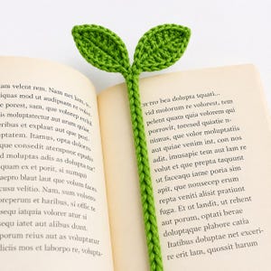 Puede incluir: Un marcapáginas de hojas verdes de ganchillo se inserta en un libro abierto. El marcapáginas presenta dos hojas en la parte superior y un tallo trenzado. Las páginas del libro están amarillentas y se ve texto.