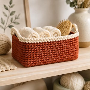 May include: A rectangular crochet basket in a deep rust-orange color with a thick cream-colored braided rim and two side handles. The basket contains three rolled cream-colored towels and a wooden hairbrush. Dimensions are 10 in long x 7 in wide x 4.5 in tall.
