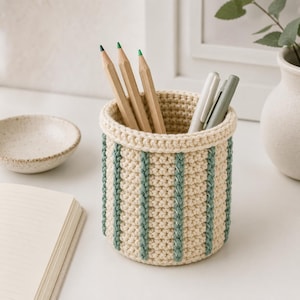 Può includere: Un portamatite cilindrico all'uncinetto con base color crema e strisce verticali in verde salvia tenue. Il bordo superiore è rifinito con un bordo spesso all'uncinetto color crema. Tre matite in legno e due penne sono all'interno del contenitore.