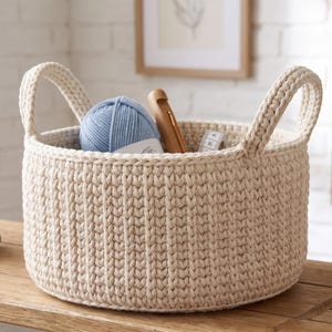May include: A round, cream-colored crocheted basket with two integrated handles. The basket contains a ball of light blue yarn, a wooden crochet hook, and a white measuring tape. The texture features a vertical ribbed stitch pattern throughout.
