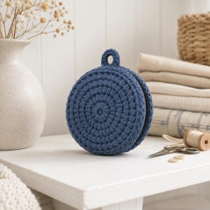 May include: A circular, dark blue crocheted pouch featuring a small loop at the top and a black zip closure along the side. The pouch is constructed with a textured, concentric stitch pattern and maintains a firm, rounded shape.