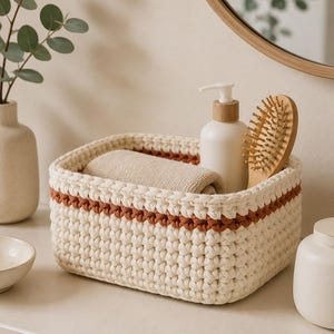 May include: A rectangular, hand-crocheted storage basket made of thick, off-white yarn with a single horizontal stripe of rust-colored yarn near the top rim. The basket contains a folded beige towel, a white pump bottle, and a wooden hairbrush.